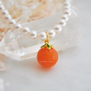 May include: A pearl necklace with a gold pendant shaped like an orange. The orange pendant is a vibrant orange color with a textured surface and green and gold accents. The necklace is on a white surface.