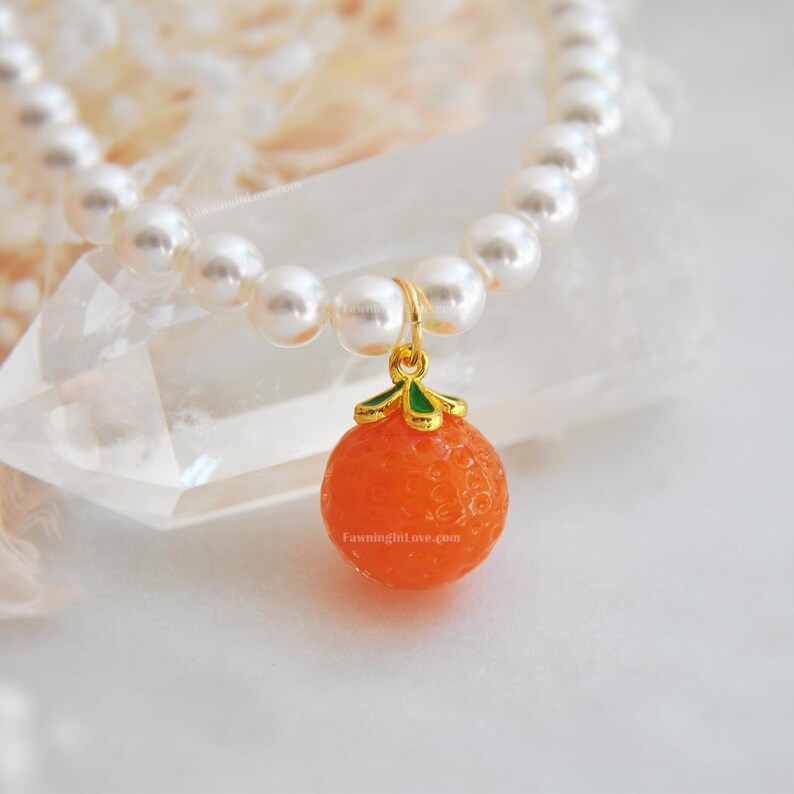 May include: A pearl necklace with a gold pendant shaped like an orange. The orange pendant is a vibrant orange color with a textured surface and green and gold accents. The necklace is on a white surface.