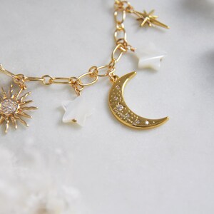 May include: Gold-toned charm bracelet featuring celestial-themed charms. The bracelet includes a sunburst charm with clear stones, a crescent moon charm with embedded crystals, and star-shaped charms. The chain is a delicate link style.