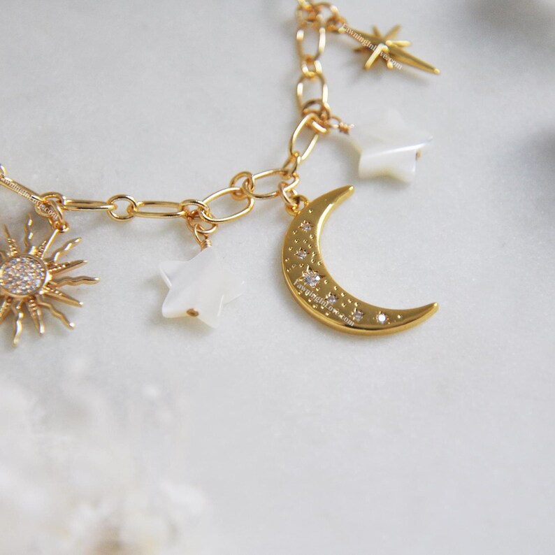 May include: Gold-toned charm bracelet featuring celestial-themed charms. The bracelet includes a sunburst charm with clear stones, a crescent moon charm with embedded crystals, and star-shaped charms. The chain is a delicate link style.
