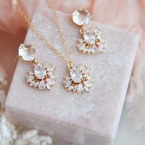 May include: A gold-toned jewelry set featuring a necklace and earrings. The necklace has a pendant with a central oval-cut clear stone surrounded by smaller clear stones. The earrings have a similar design, with a large square-cut stone at the top.