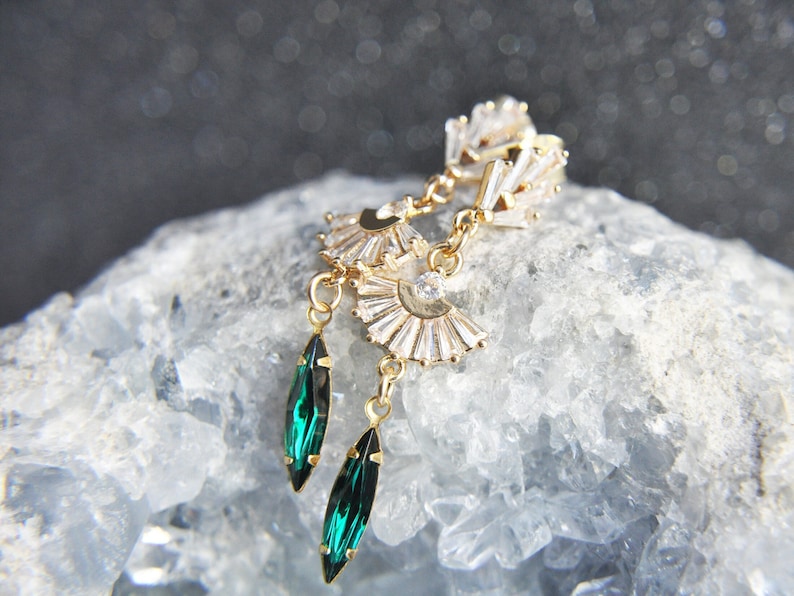 May include: Gold-toned earrings with a fan-shaped design and emerald green teardrop-shaped pendants. The earrings feature clear, rectangular and round crystals. The earrings are displayed on a light blue, crystalline surface.