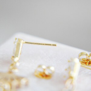 May include: Close-up of gold-colored stud earrings with a post and backing. The earrings have a small, clear gemstone design. The background is a soft, neutral color.