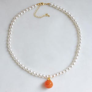 May include: A delicate pearl necklace with a gold-toned clasp and a small, orange, textured pendant. The necklace features a strand of small, round, white pearls. The pendant is a spherical shape with a textured surface.