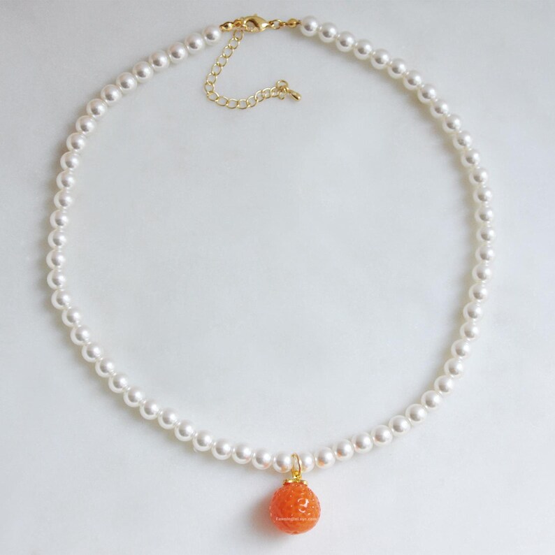 May include: A delicate pearl necklace with a gold-toned clasp and a small, orange, textured pendant. The necklace features a strand of small, round, white pearls. The pendant is a spherical shape with a textured surface.