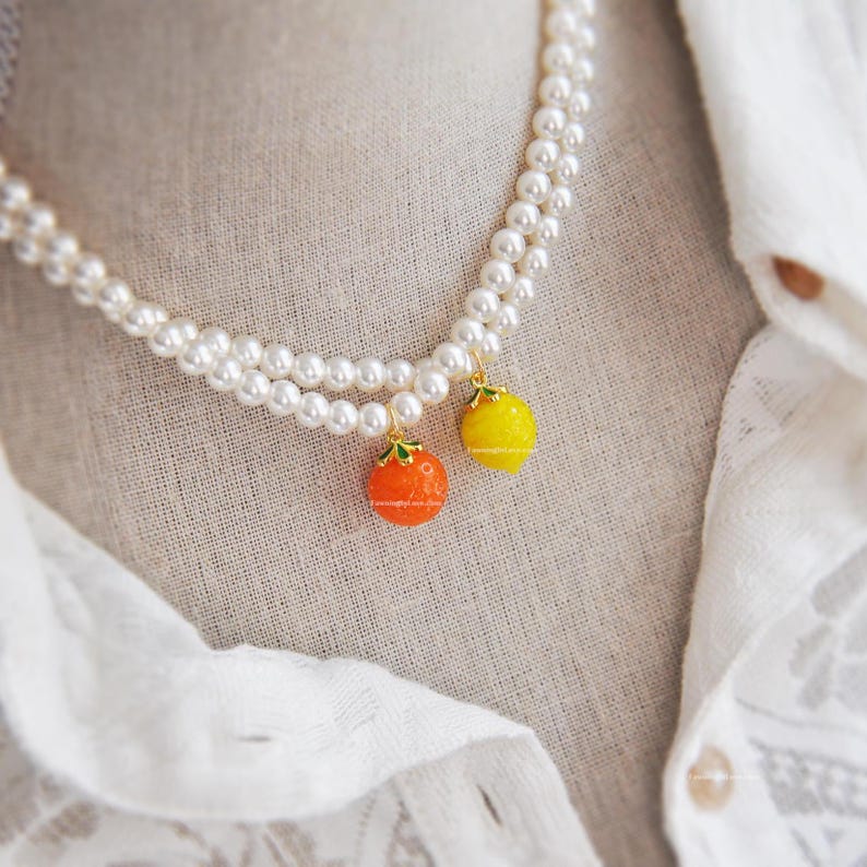 May include: A double-strand pearl necklace with two fruit-shaped pendants. One pendant is orange, the other is yellow, both with green accents. The necklace is displayed on a white shirt.
