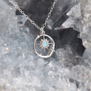 Puede incluir: Un collar plateado con un colgante celestial. El colgante presenta una luna creciente, un dise&ntilde;o de estrella y una &oacute;palo ovalada central. La cadena fina del collar resalta una mezcla de elementos celestiales y naturales.