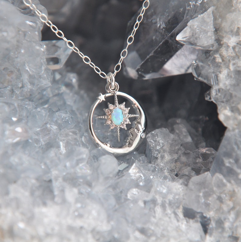 Puede incluir: Un collar plateado con un colgante celestial. El colgante presenta una luna creciente, un dise&ntilde;o de estrella y una &oacute;palo ovalada central. La cadena fina del collar resalta una mezcla de elementos celestiales y naturales.