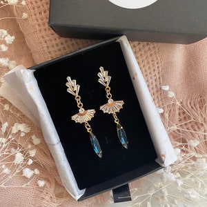 May include: A pair of gold-toned earrings with a fan-shaped design and clear crystal accents. Each earring features a teardrop-shaped, dark blue crystal pendant. The earrings are displayed in a black jewelry box with the brand name "Fawning in Love Jewelry".