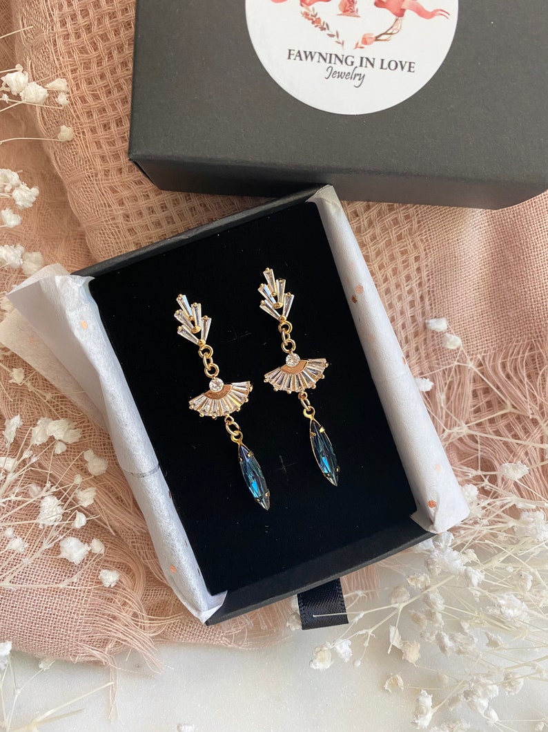 May include: A pair of gold-toned earrings with a fan-shaped design and clear crystal accents. Each earring features a teardrop-shaped, dark blue crystal pendant. The earrings are displayed in a black jewelry box with the brand name "Fawning in Love Jewelry".