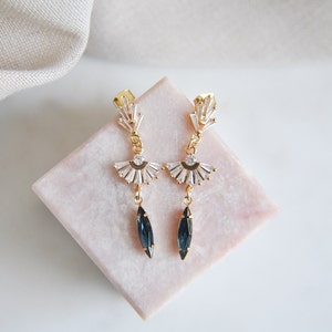 May include: A pair of gold-toned earrings with a fan-shaped design, adorned with clear crystals and a dark blue teardrop-shaped stone. The earrings are displayed on a pink square surface.
