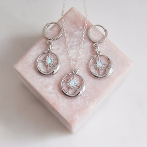 Puede incluir: Un conjunto de joyas plateadas que incluye un collar y pendientes de aro. Cada pieza presenta un dise&ntilde;o celestial con un motivo de luna y estrella, y una piedra central similar al &oacute;palo. El conjunto se exhibe sobre una superficie geom&eacute;trica rosa.