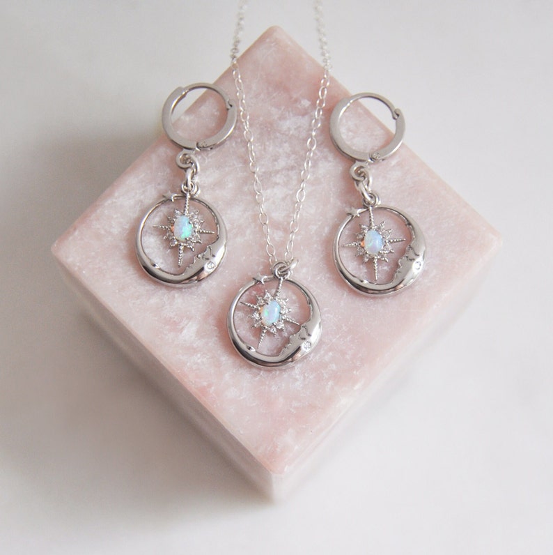 Puede incluir: Un conjunto de joyas plateadas que incluye un collar y pendientes de aro. Cada pieza presenta un dise&ntilde;o celestial con un motivo de luna y estrella, y una piedra central similar al &oacute;palo. El conjunto se exhibe sobre una superficie geom&eacute;trica rosa.