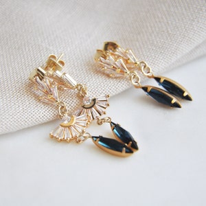 May include: Gold-toned dangle earrings with a fan-shaped design, featuring clear baguette-cut crystals and dark blue teardrop-shaped gems. The earrings have a post-back closure and are displayed on a neutral fabric background.