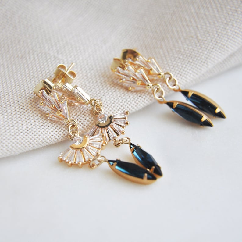 May include: Gold-toned dangle earrings with a fan-shaped design, featuring clear baguette-cut crystals and dark blue teardrop-shaped gems. The earrings have a post-back closure and are displayed on a neutral fabric background.