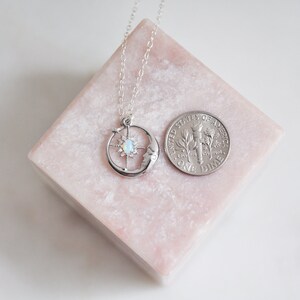 Puede incluir: Un collar plateado con un dise&ntilde;o celestial que presenta una luna creciente, una estrella y una piedra central similar al &oacute;palo. El collar se muestra en un bloque rosa junto a una moneda para referencia de tama&ntilde;o.