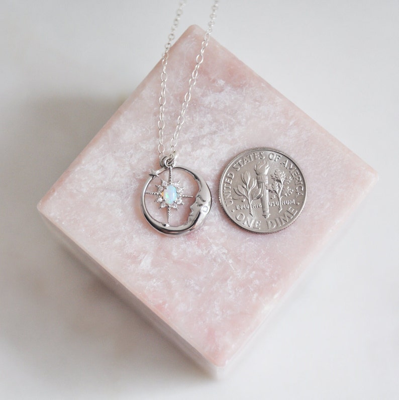 Puede incluir: Un collar plateado con un dise&ntilde;o celestial que presenta una luna creciente, una estrella y una piedra central similar al &oacute;palo. El collar se muestra en un bloque rosa junto a una moneda para referencia de tama&ntilde;o.
