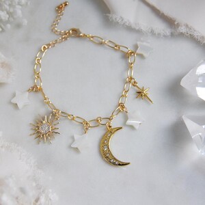 May include: Gold-toned bracelet featuring celestial charms. The bracelet includes a sunburst pendant with a central cluster of small stones, a crescent moon pendant with embedded stones, and star-shaped charms. The chain is a linked design.