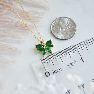 Puede incluir: Un delicado collar dorado con un colgante en forma de lazo adornado con brillantes piedras preciosas verdes. El colgante mide aproximadamente 1,3 cm de ancho. Se incluye una regla y una moneda como referencia de tama&ntilde;o. El collar se muestra sobre una superficie blanca.