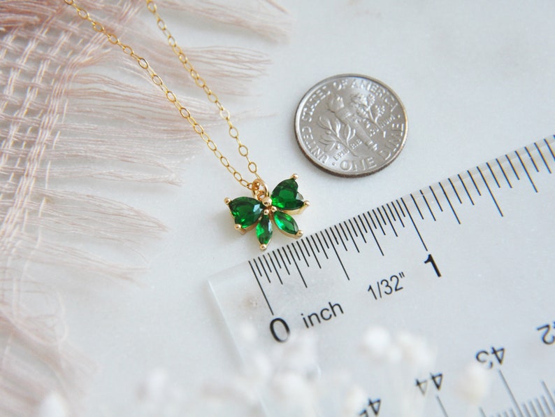Puede incluir: Un delicado collar dorado con un colgante en forma de lazo adornado con brillantes piedras preciosas verdes. El colgante mide aproximadamente 1,3 cm de ancho. Se incluye una regla y una moneda como referencia de tama&ntilde;o. El collar se muestra sobre una superficie blanca.