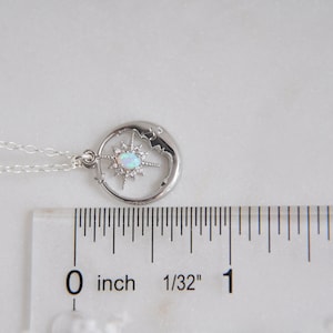 Puede incluir: Un collar plateado con un colgante celestial. El colgante presenta un dise&ntilde;o de media luna con una estrella en el centro, adornado con una piedra blanca similar al &oacute;palo y peque&ntilde;os cristales transparentes. La cadena es delicada. El colgante mide unos 2,5 cm de di&aacute;metro.
