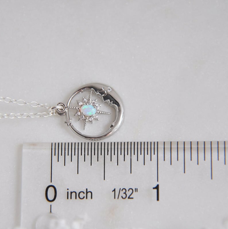 Puede incluir: Un collar plateado con un colgante celestial. El colgante presenta un dise&ntilde;o de media luna con una estrella en el centro, adornado con una piedra blanca similar al &oacute;palo y peque&ntilde;os cristales transparentes. La cadena es delicada. El colgante mide unos 2,5 cm de di&aacute;metro.