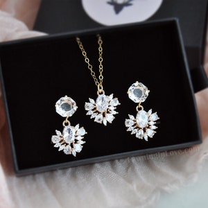 May include: A gold-toned jewelry set featuring a necklace and matching earrings, presented in a black box. The set includes a pendant necklace and drop earrings, each adorned with clear, sparkling crystals. The jewelry is designed with a floral-inspired motif.