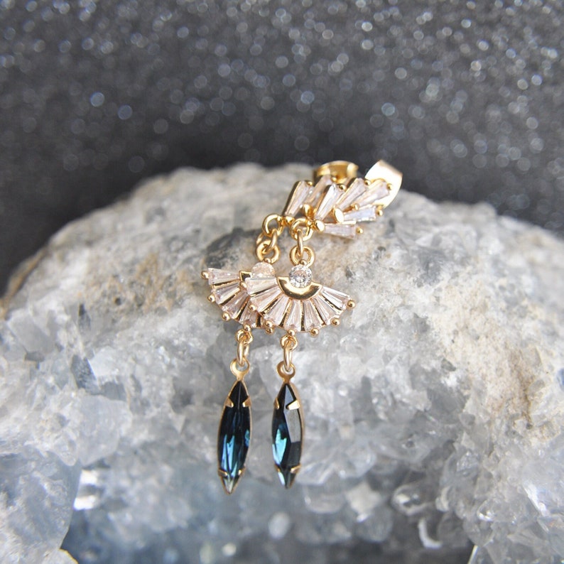 May include: Gold-toned earring with a fan-shaped design of clear crystals and two dangling, dark blue, teardrop-shaped crystals. The earring is set against a light blue, textured background.