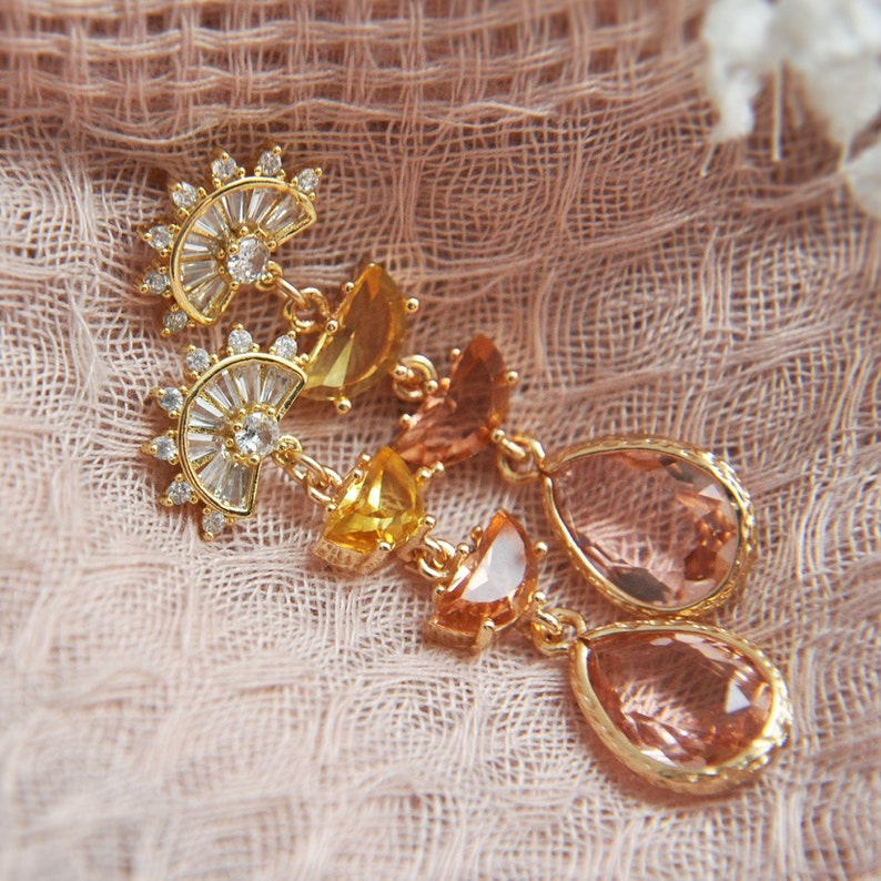 May include: Gold-toned earrings with a fan-shaped top, clear and yellow crystals, and teardrop-shaped peach-colored gems. The earrings are set against a soft, textured pink background.