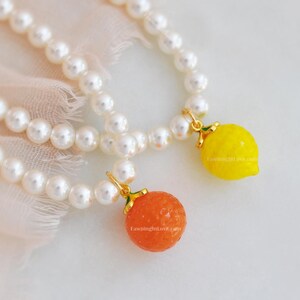 May include: Two pearl necklaces with fruit charms. One necklace has a yellow lemon charm, and the other has an orange charm. The charms are attached with gold-colored findings. The website "FawningInLove.com" is printed on the charms.