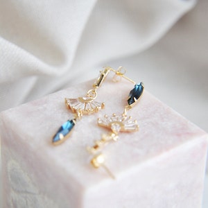 May include: Gold-toned earrings with a fan-shaped design and blue gemstone accents. The earrings feature a post-back closure and are displayed on a pink marble surface. The gemstones are a deep blue color.