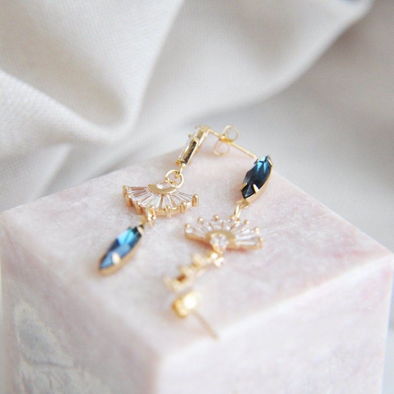 May include: Gold-toned earrings with a fan-shaped design and blue gemstone accents. The earrings feature a post-back closure and are displayed on a pink marble surface. The gemstones are a deep blue color.