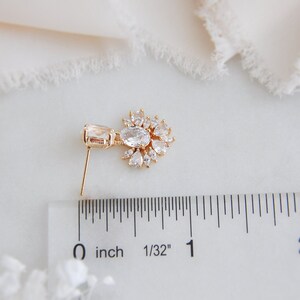 May include: A gold-colored earring with a rectangular clear stone and a cluster of clear, teardrop-shaped stones. The earring is approximately 1 inch long, as measured by the ruler in the image. The earring has a post for pierced ears.