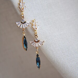 May include: A pair of gold-toned earrings with a fan-shaped design and clear crystals. Each earring features a dangling, dark blue, marquise-cut crystal. The earrings are displayed against a neutral background, showcasing their elegant design.