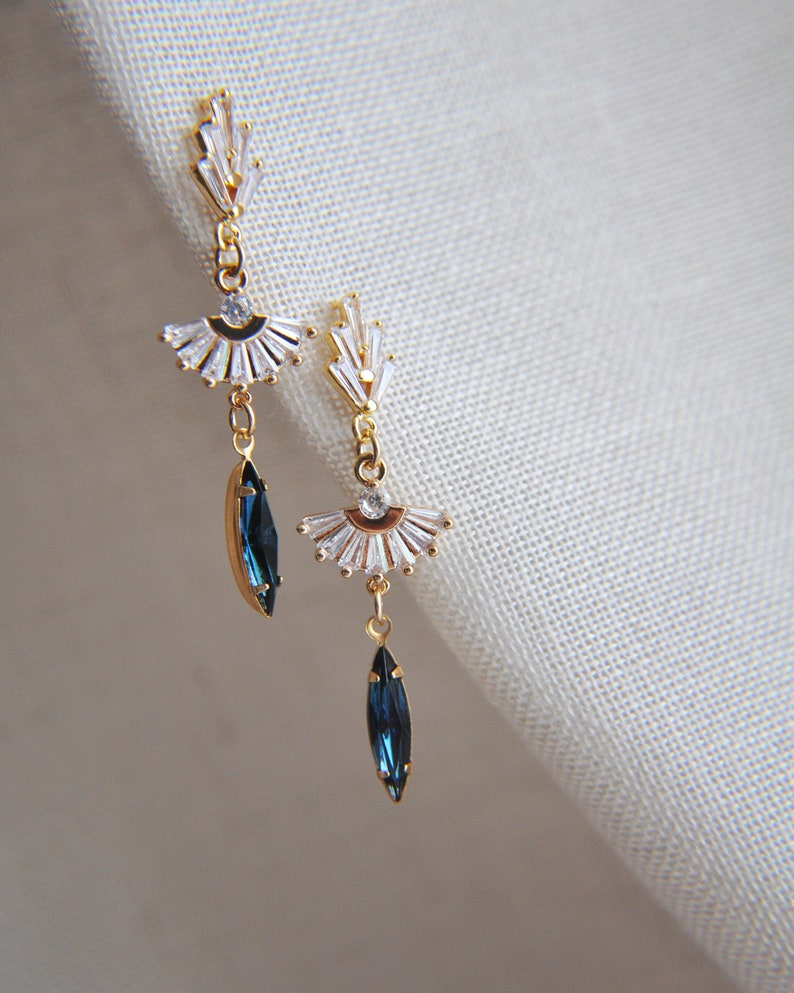 May include: A pair of gold-toned earrings with a fan-shaped design and clear crystals. Each earring features a dangling, dark blue, marquise-cut crystal. The earrings are displayed against a neutral background, showcasing their elegant design.