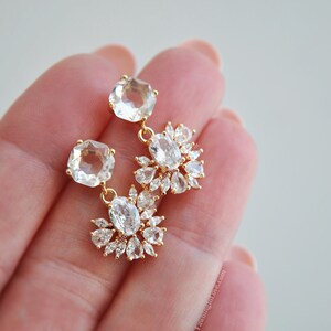 May include: Gold-toned drop earrings featuring clear, faceted gemstones. Each earring has a square stone at the top, connected to a cluster of oval and teardrop-shaped stones. The earrings are held in a hand.