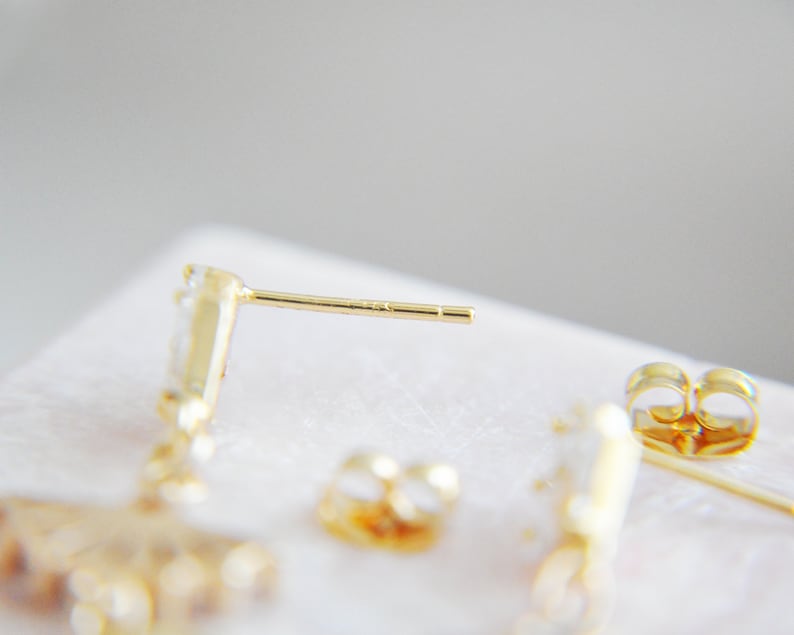 May include: Close-up of gold-colored stud earrings with a post and backing. The earrings have a small, clear gemstone design. The background is a soft, neutral color.