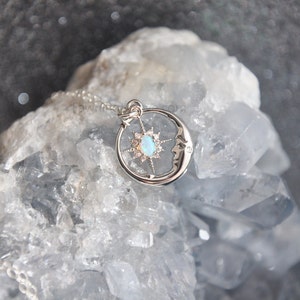 Puede incluir: Un collar con colgante plateado con un dise&ntilde;o celestial. El colgante incluye una luna creciente con una cara, una estrella y una piedra central similar a un &oacute;palo. El collar se muestra sobre una superficie de cristal azul claro.