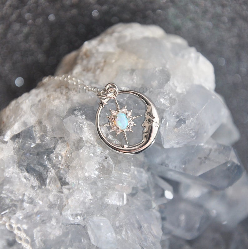Puede incluir: Un collar con colgante plateado con un dise&ntilde;o celestial. El colgante incluye una luna creciente con una cara, una estrella y una piedra central similar a un &oacute;palo. El collar se muestra sobre una superficie de cristal azul claro.