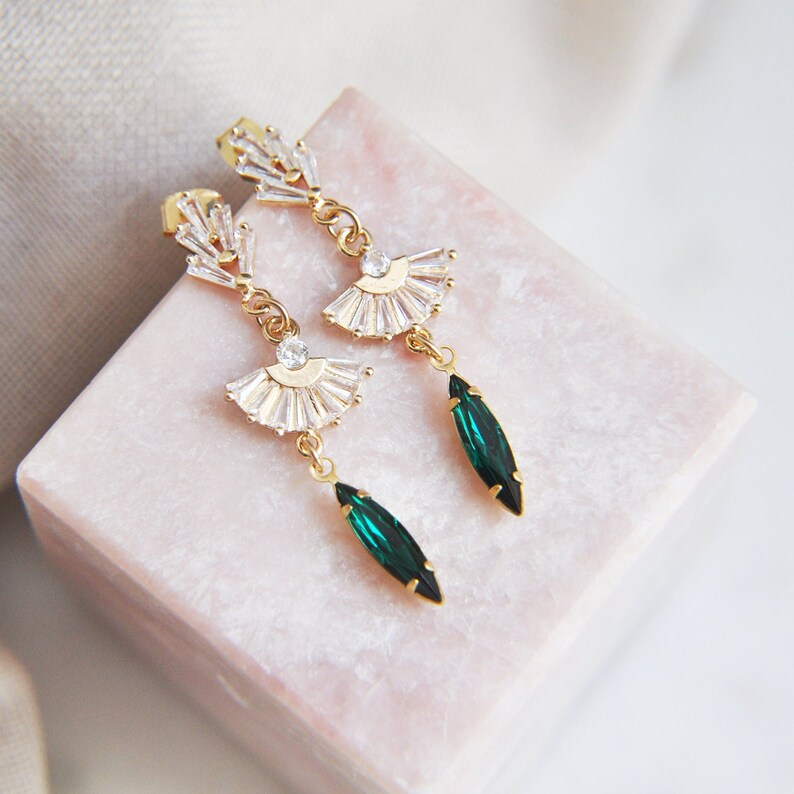 May include: A pair of gold-toned earrings with a fan-shaped design of clear crystals and a dangling emerald green teardrop-shaped crystal. The earrings are displayed on a pink surface.