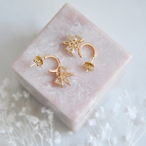 Puede incluir: Un par de pendientes de aro dorados con un dise&ntilde;o floral. Cada pendiente presenta un peque&ntilde;o colgante de flor con detalles de piedras preciosas transparentes. Los pendientes se exhiben sobre un bloque cuadrado rosa.