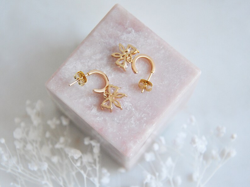 Puede incluir: Un par de pendientes de aro dorados con un dise&ntilde;o floral. Cada pendiente presenta un peque&ntilde;o colgante de flor con detalles de piedras preciosas transparentes. Los pendientes se exhiben sobre un bloque cuadrado rosa.
