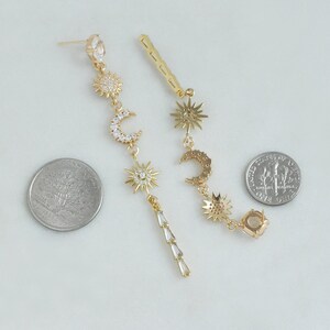 May include: Gold-toned celestial earrings featuring sun and moon charms with clear crystal accents. One earring has a dangling design with a series of baguette-cut crystals. The earrings are shown next to a quarter and a dime for size reference.