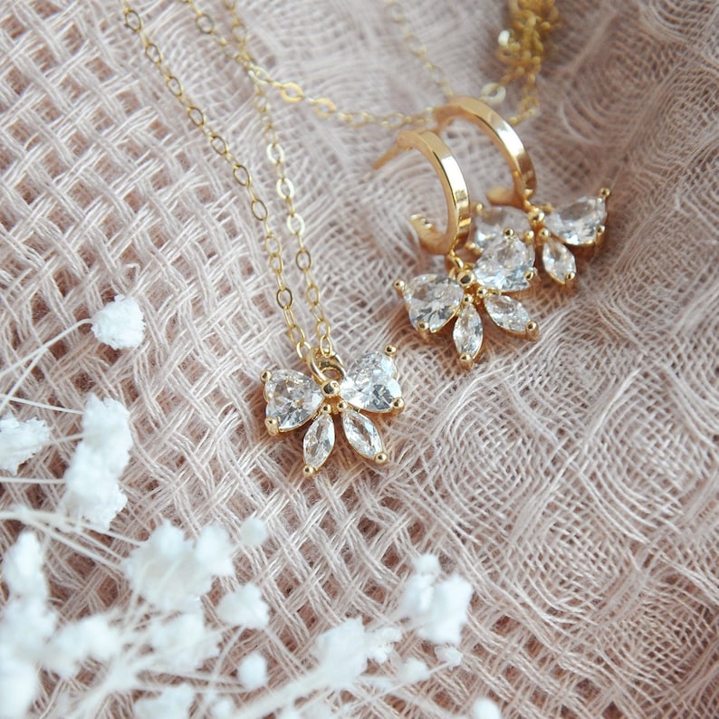 K&ouml;nnte beinhalten: Ein goldfarbenes Schmuckset mit Halskette und Ohrringen. Die Halskette hat eine feine Kette mit einem schleifenf&ouml;rmigen Anh&auml;nger, der mit klaren Kristallen verziert ist. Die Ohrringe sind im Creolen-Stil gehalten, ebenfalls mit schleifenf&ouml;rmigen Kristallanh&auml;ngern. Das Schmuckset wird auf einer strukturierten, hellrosa Oberfl&auml;che pr&auml;sentiert.