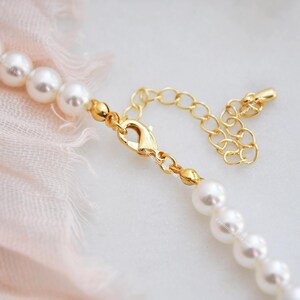May include: Close-up of a pearl necklace with a gold-colored clasp and extender chain. The necklace features multiple white, round pearls. The clasp is a lobster claw style. The background is a soft pink fabric.