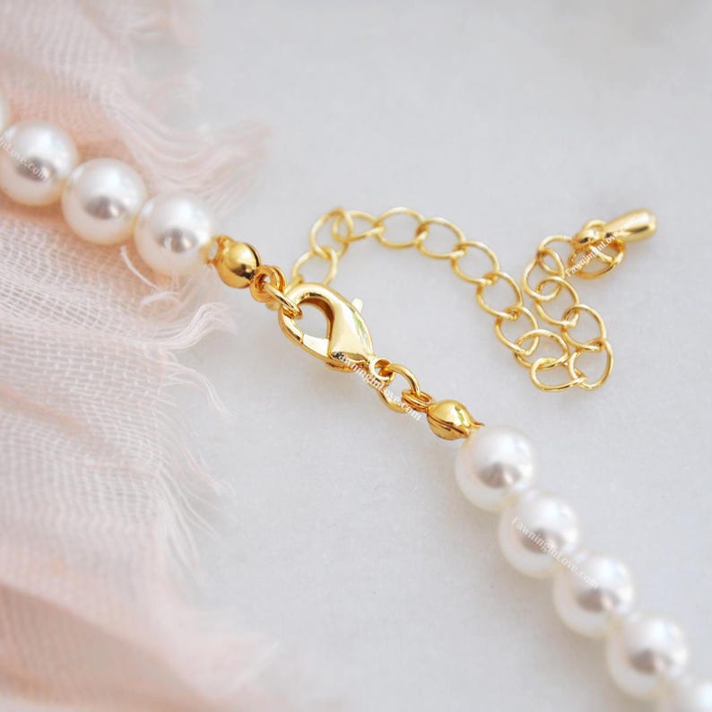 May include: Close-up of a pearl necklace with a gold-colored clasp and extender chain. The necklace features multiple white, round pearls. The clasp is a lobster claw style. The background is a soft pink fabric.