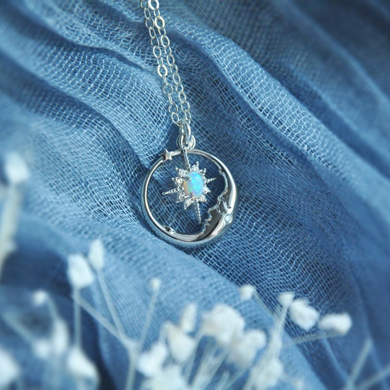 Puede incluir: Un collar plateado con un colgante celestial. El colgante presenta una estrella con una &oacute;palo central, una luna creciente con una cara y una peque&ntilde;a estrella. El collar cuelga de una delicada cadena sobre un fondo de tela azul.