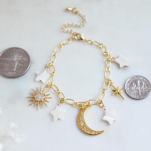 May include: Gold-toned chain bracelet with celestial charms. The bracelet features a sunburst charm with a central crystal, a crescent moon charm, and several star-shaped charms. The bracelet is shown with a quarter and a dime for size reference.