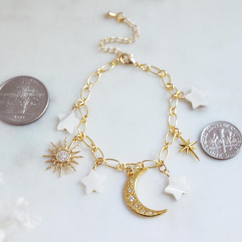 May include: Gold-toned chain bracelet with celestial charms. The bracelet features a sunburst charm with a central crystal, a crescent moon charm, and several star-shaped charms. The bracelet is shown with a quarter and a dime for size reference.