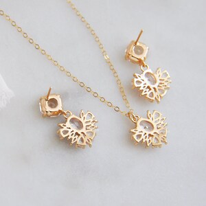 May include: A gold-colored jewelry set featuring a necklace and a pair of earrings. Each piece has a square-shaped top with a dangling floral design, all adorned with clear gemstones. The set is displayed on a white surface.